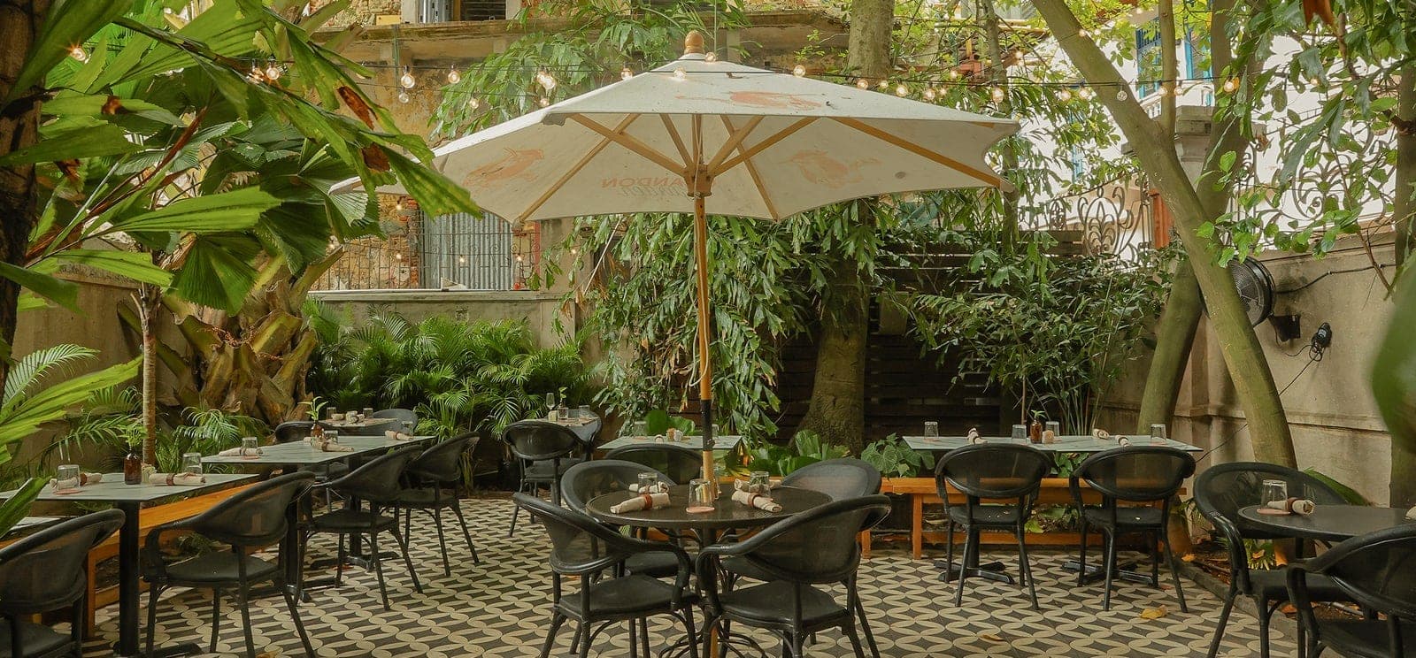 Patio jardín frondoso con mesas bajo sombrilla y luces colgantes en Mahalo Cocina y Jardin, Casco Viejo, Ciudad de Panamá